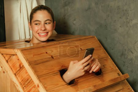 Photo for A portrait of gorgeous caucasian woman playing her mobile phone while using wooden sauna cabinet in warm tone. Attractive female with beautiful skin taking a photo. Gray background. Tranquility - Royalty Free Image