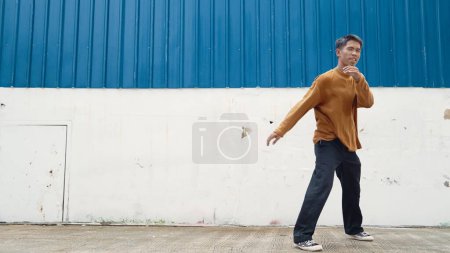Foto de El hombre hispano estira los brazos y baila en la calle bailando frente a la pared. Motion shot de bailarina con estilo o coreógrafa en traje casual que practica el baile en estilo hip hop. Deporte al aire libre 2024. Endeavor. - Imagen libre de derechos