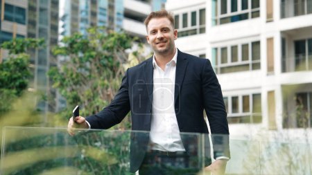 Photo for Skilled caucasian business man looking at camera while standing at rooftop. Professional investor smiling with confident while wearing formal suit at modern building in green city. Lifestyle. Urbane - Royalty Free Image