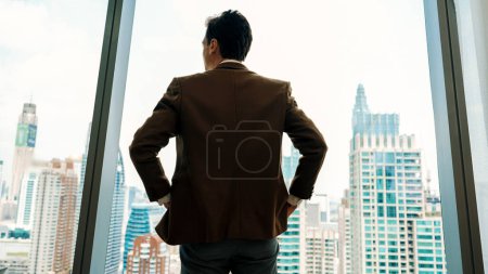 Foto de Vista posterior ambicioso hombre de negocios de pie en la oficina ornamentada mirando por la ventana al horizonte del paisaje urbano. La determinación y la ambición empresarial impulsan la carrera empresarial hacia un futuro brillante - Imagen libre de derechos