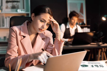 Photo for Focusing on sitting woman coworker over thinking campaign writing on paperwork and laptop on working desk while man leaning at desk waiting email at late night time with blurry background. Postulate. - Royalty Free Image