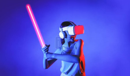 Téléchargez les photos : Femme intelligente portant un casque VR connectant la métaverse, future technologie de la communauté cyberspatiale. Femme élégante tenant fermement sabre laser sérieusement tout en jouant jeu de combat d'épée. Hallucination. - en image libre de droit