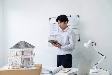 Professionelle gut aussehende asiatische Projektmanager mit Tablet, um Hausmodell zu überprüfen, während Hausmodell Struktur sorgfältig im Büro mit Bauplan hinter gehängt überprüfen. Kreatives Design. Makellos.