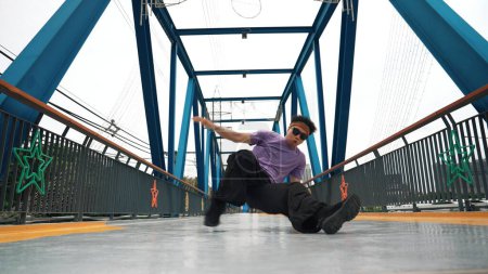 Téléchargez les photos : Hipster effectuer b-boy pas et la danse de rue au pont avec fond. Le danseur professionnel de pause porte un tissu élégant et regarde la caméra tout en posant à la caméra. Sport de plein air 2024. Brillamment. - en image libre de droit