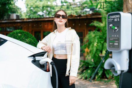 Photo for Young woman and sustainable urban commute with EV electric car recharging at outdoor cafe in springtime garden, green city sustainability and environmental friendly EV car. Expedient - Royalty Free Image