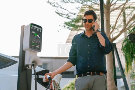 Photo for Young man recharge EV electric vehicle at green city park parking lot while talking on phone. Sustainable urban lifestyle for environment friendly EV car with battery charging station. Expedient - Royalty Free Image