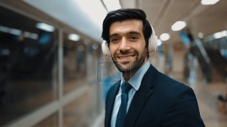 Photo for Closeup of smart business man wearing headphone and listening music while standing at train station. Professional executive manager looking at camera while waiting for train or subway. Exultant. - Royalty Free Image