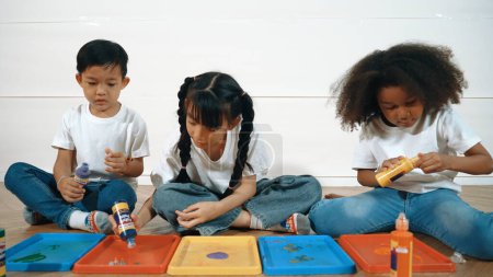Téléchargez les photos : Groupe de jeunes étudiants diversifiés mélangeant les couleurs tout en étant assis ensemble. Heureux enfants multiculturels préparés mélangés à jouer et à créer de nouvelles œuvres d'art à la leçon d'art. Concept d'activité créative. Erudition. - en image libre de droit