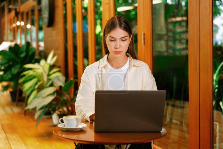 Mujer joven que trabaja en el ordenador portátil en el jardín de la cafetería al aire libre durante la primavera, disfrutando de ambiente sereno en la cafetería. Freelancer nómada digital o estudiante universitario trabajando remotamente o blogueando. Expediente