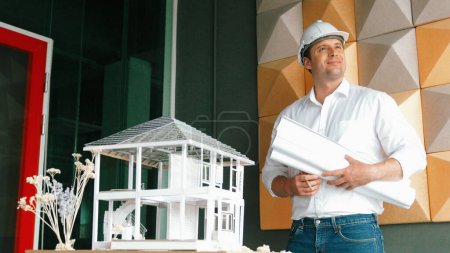 Photo for Portrait of professional project manager holding project plan with house model at table. Professional architect engineer wearing safety helmet while standing posing with confident. Manipulator. - Royalty Free Image
