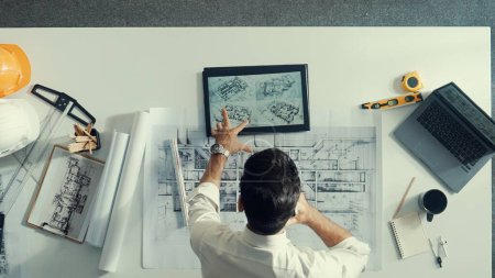Photo for Top view of skilled architect looking at project plan at tablet. Aerial view of civil engineer planning while checking building blueprint at meeting room with safety helmet and laptop. Alimentation. - Royalty Free Image