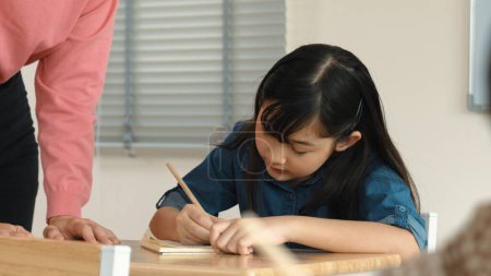 Chica asiática escribiendo o tomando una nota mientras el profesor revisa el trabajo en clase. Maestra caucásica ayudar a estudiante feliz haciendo prueba o trabajo de clase, mientras que diversos estudiantes estudian en la lección de ciencias. Educación. Pedagogía.