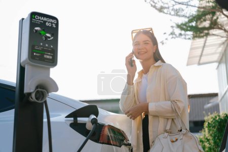Photo for Young woman holding shopping bag talking on the phone while recharging EV car battery from charging station at city mall parking lot. Modern woman go shopping by eco car. Expedient - Royalty Free Image
