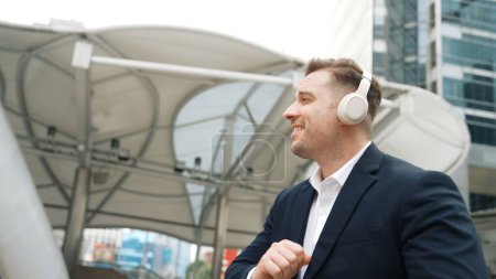 Glücklicher Geschäftsmann, der über Kopfhörer Entspannungsmusik hört und sich mit Musik bewegt, während er mit lebhafter Stimmung auf der Straße in der Großstadt spaziert. Manager tragen Headset und hören entspannt Reimen zu. Urbane Pflanzen.