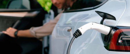 Photo for Young man recharge electric cars battery from charging station in outdoor green city park. Rechargeable EV car for sustainable environmental friendly urban travel. Panorama Expedient - Royalty Free Image