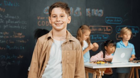 Photo for Smart boy smiling at camera while diverse friend working or learning engineering code or prompt in STEM technology classroom. Cute student standing at camera while children using laptop. Erudition. - Royalty Free Image