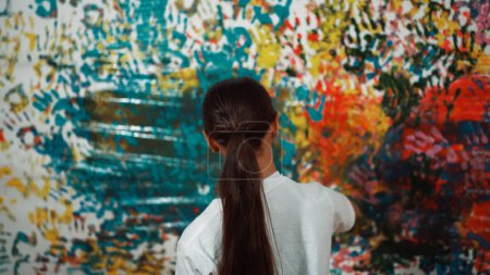 Photo for Young happy girl holding the color tray and paint colorful stained wall to express idea and enhance imagination with hand print. Caucasian girl stamp hand to decorate room in art lesson. Edification. - Royalty Free Image