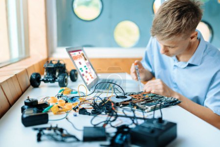 Photo for Schoolboy in blue shirt connect parts of robotics vehicle with motherboard carefully in STEM class. On table put laptop, controller, electric wire, battery charger, and robotic vehicle. Edification. - Royalty Free Image