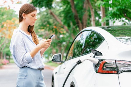 Photo for Young woman recharge EV electric vehicle battery from EV charging station and using smartphone online banking to pay for electricity in city park. Eco friendly vehicle travel with EV car. Exalt - Royalty Free Image