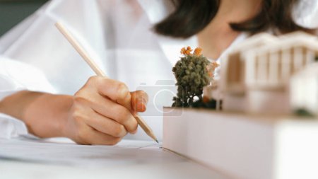 Photo for Closeup of young beautiful architect engineer drawing blueprint while looking at architectural model to analyze house construction. Project manager check house model during taking a note. Manipulator. - Royalty Free Image