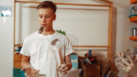 Photo for Smiling caucasian boy looking at camera and throw clay while stand in front of blackboard. Happy multicultural student hold dough while look at camera in art lesson. Creative activity. Edification. - Royalty Free Image