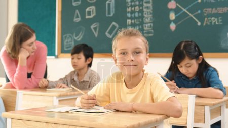 Téléchargez les photos : Enfant intelligent caucasien souriant à la caméra tout en faisant du travail en classe tout en étudiant multiculturel heureux faire des tests ou note d'écriture en classe au primaire. Concept d'éducation. Pédagogie. - en image libre de droit