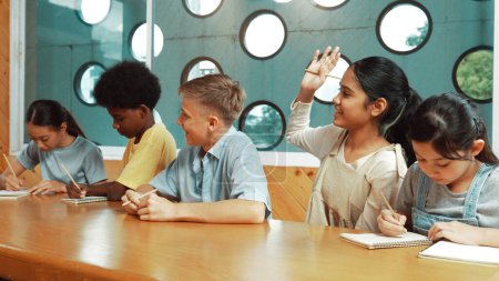 Photo for Diverse student writing answer while girl raised hand to ask question, voting, volunteering, asking .answering question at table in classroom. Multicultural children working together. Edification. - Royalty Free Image