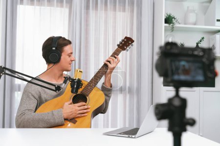 Photo for Host channel in smart singer recording by camera, playing guitar along singing, broadcasting on social media channel, wearing headphones to record video streamer at white modern studio. Pecuniary. - Royalty Free Image