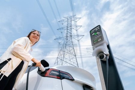 Woman recharge EV electric car battery at charging station connected to electrical power grid tower on sky background as electrical industry for eco friendly vehicle utilization. Expedient