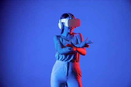Foto de Mujer inteligente de pie con auriculares VR que conectan metaverse, tecnología futura de la comunidad del ciberespacio. Mujer elegante con las manos al mando pistola virtual jugando seriamente juegos de disparos. Alucinaciones. - Imagen libre de derechos
