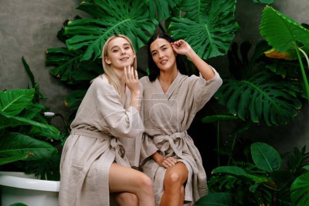Tropischer und exotischer Kurgarten mit Badewanne im modernen Hotel oder Resort mit zwei jungen Frauen im Bademantel, die inmitten üppig grüner Laubkulisse ihren Freizeit- und Wellness-Lifestyle genießen. Glückseligkeit