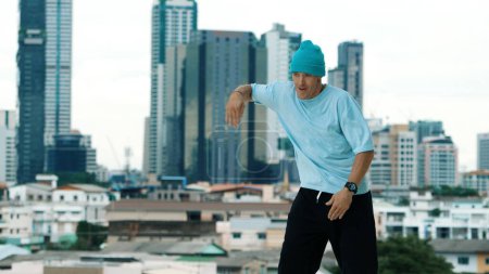 Téléchargez les photos : Danseur B-boy caucasien qualifié pratiquant la danse de rue sur le toit avec des sites urbains ou urbains. Prise de vue en mouvement d'un jeune homme exécutant une danse de rue en posant sur gel. Sport de plein air 2024. Endeavor. - en image libre de droit