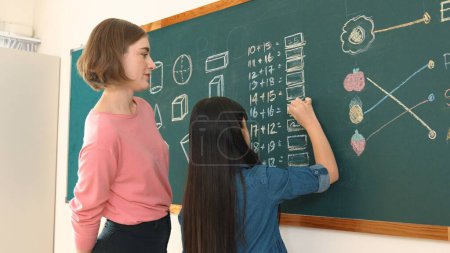 Photo for Smart caucasian girl raised hand and volunteer to write answer on blackboard. Teacher ask for volunteer to answer question while happy cute student answer question and walk back to her seat. Pedagogy. - Royalty Free Image