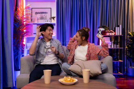 Photo for Couple gamer raising fist up holding joysticks be winner together complete level sitting sofa playing video game with front snack and drinks at neon light color living room at modern home. Infobahn. - Royalty Free Image