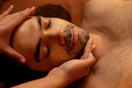 Photo for Caucasian man enjoying relaxing anti-stress head massage and pampering facial beauty skin recreation leisure in warm candle lighting ambient salon spa in luxury resort or hotel. Quiescent - Royalty Free Image