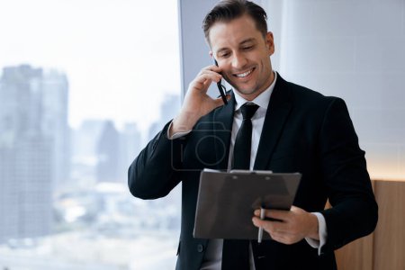 Photo for Corporate skilled businessman talking phone and looking at data while standing near window shows skyscraper view. Serious busy manager working by phone. Modern workplace, office. Business. Ornamented. - Royalty Free Image