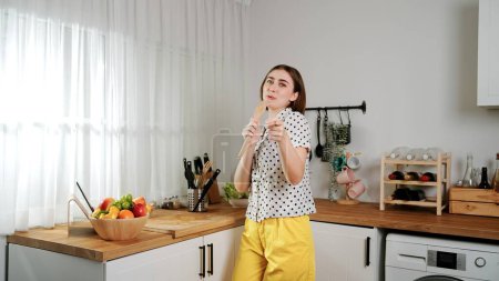 Photo for Energetic caucasian mother singing by holding ladle at modern kitchen while cooking and making salad for breakfast. Happy house keeper moving to music while prepare for making healthy food. Pedagogy. - Royalty Free Image
