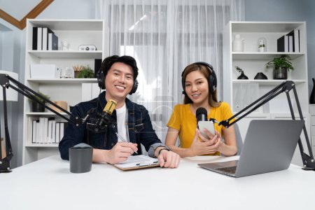 Photo for Smiling radio influencer hosts wearing headphones looking camera to pose on social online live streaming with talking topic script note and laptop to listeners at morning at studio record. Infobahn. - Royalty Free Image