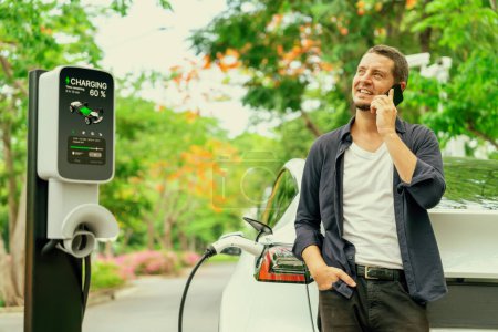 Ein Mann telefoniert mit dem Smartphone, während er im Urlaub im Nationalpark oder im herbstlichen Wald den Akku eines Elektroautos an einer Ladestation auflädt. Erhöhen