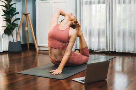 Téléchargez les photos : Femme flexible et dextérité en tenue de sport faisant position de yoga en posture de méditation sur tapis d'exercice à la maison. Gaieté saine séance de formation en ligne de yoga à domicile avec l'esprit paisible et la sérénité. - en image libre de droit