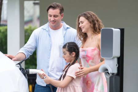 Photo for Happy little young girl learn about eco-friendly and energy sustainability as she help her family recharge electric vehicle from home EV charging station. EV car and modern family concept. Synchronos - Royalty Free Image