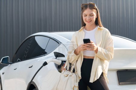 Photo for Young woman holding shopping bag and use smartphone to pay for electricity for recharging EV car battery from charging station at city mall parking lot. Modern woman go shopping by eco car. Expedient - Royalty Free Image