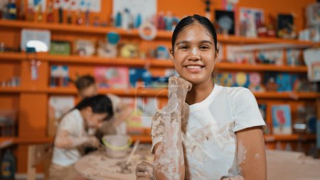 Photo for Happy caucasian girl pose at camera while diverse children modeling clay behind. Cute student wearing dirty shirt while looking at camera at workshop in art lesson. Blurring background. Edification. - Royalty Free Image