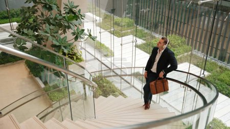 Top view of caucasian businessman listening relaxing music wearing headphone while standing at spiral stair cases at modern building. Project manager enjoy listening song while move to rhyme. Urbane.