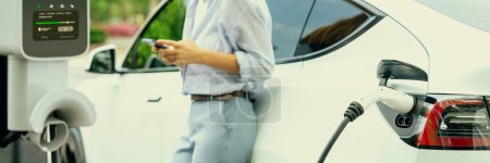 Photo for Young woman using smartphone online banking application to pay for electric car battery charging from EV charging station during vacation road trip at national park or summer forest. Panorama Exalt - Royalty Free Image