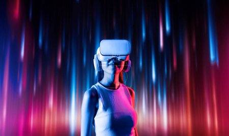 Photo for Smart female standing surrounded by neon light wearing VR headset connecting metaverse, future cyberspace community technology. Elegant woman looking faraway and smiling satisfactorily. Hallucination. - Royalty Free Image