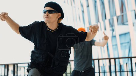 Asian hipster looking at camera while pose at camera while friend dancing behind at sky scrapper. Group of street dancer perform break dancing while looking at camera. Outdoor sport 2024. Endeavor.
