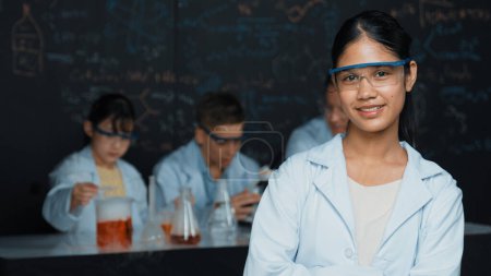 Photo for Girl looking at camera with arm folded while people doing experiment at laboratory. Cute student standing blackboard with chemical theory with blurring background at STEM science class. Edification. - Royalty Free Image