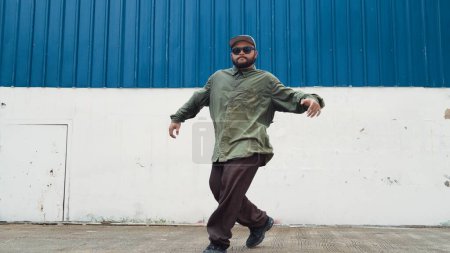 Hipster dancing b-boy foot step at street with blue wall. Asian break dancer practice street dance while stretch arms. Hip hop choreographer perform energetic dance. Outdoor sport 2024. Endeavor.