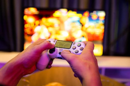 Close up one hands man player of videospiel on TV mit Joystick-Steuerung mit verschwommenen Kampf-Gaming-Bildschirm mit Snack und Getränken bei dekorativer mit Neon-Blaulicht im gemütlichen Wohnzimmer. Postulat.